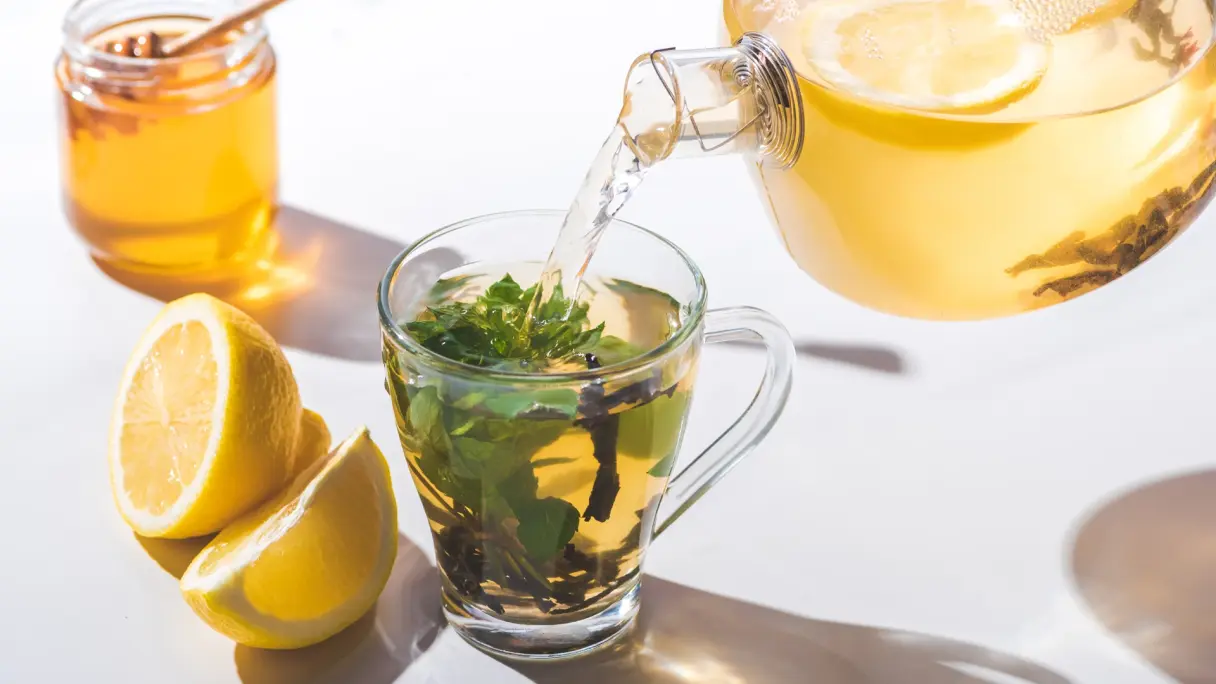 Aus einer Glaskanne wird Tee in eine gläserne Tasse gegossen. Der Tee hat eine gelbliche Farbe. In der Tasse befinden sich frische Kräuter. Daneben liegt eine aufgeschnittene Zitrone.