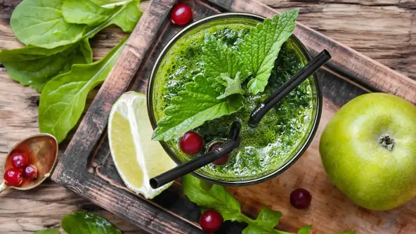 Obenansicht auf einen grünen Saft in einem Glas mit zwei Strohhalmen. Daneben liegen ein grüner Apfel, Salatblätter, ein Limettenschnitz und ein paar Johannisbeeren.