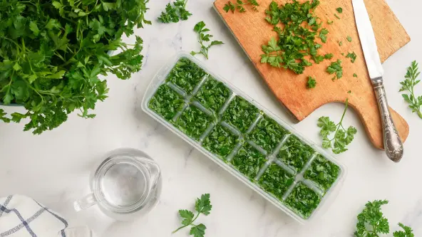 Gefrorene Kräuter zum Kochen in der Draufsicht. Frische Bio-Petersilie und die Vorbereitung von Petersilieneiswürfeln auf einem Marmorhintergrund. Daneben ist eine große Schale Petersilie sowie ein Holzschneidebrett mit Messer und ein Krug Wasser zu sehen.