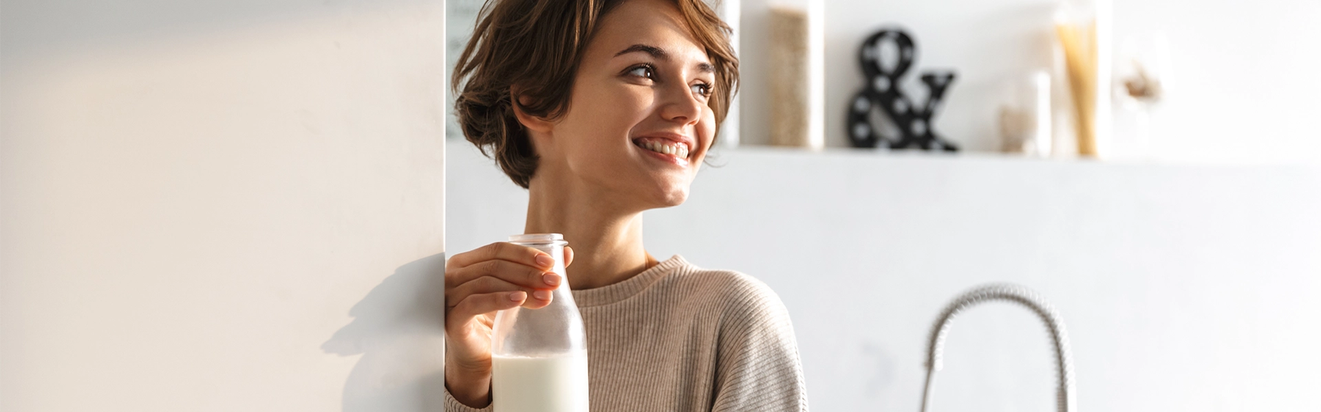Person öffnet Kühlschranktür und hält eine Milchflasche in der anderen Hand; helle, moderne Küche im Hintergrund.