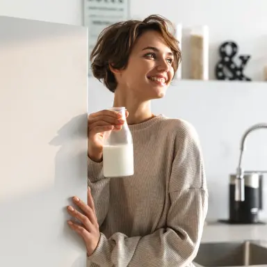Person öffnet Kühlschranktür und hält eine Milchflasche in der anderen Hand; helle, moderne Küche im Hintergrund.