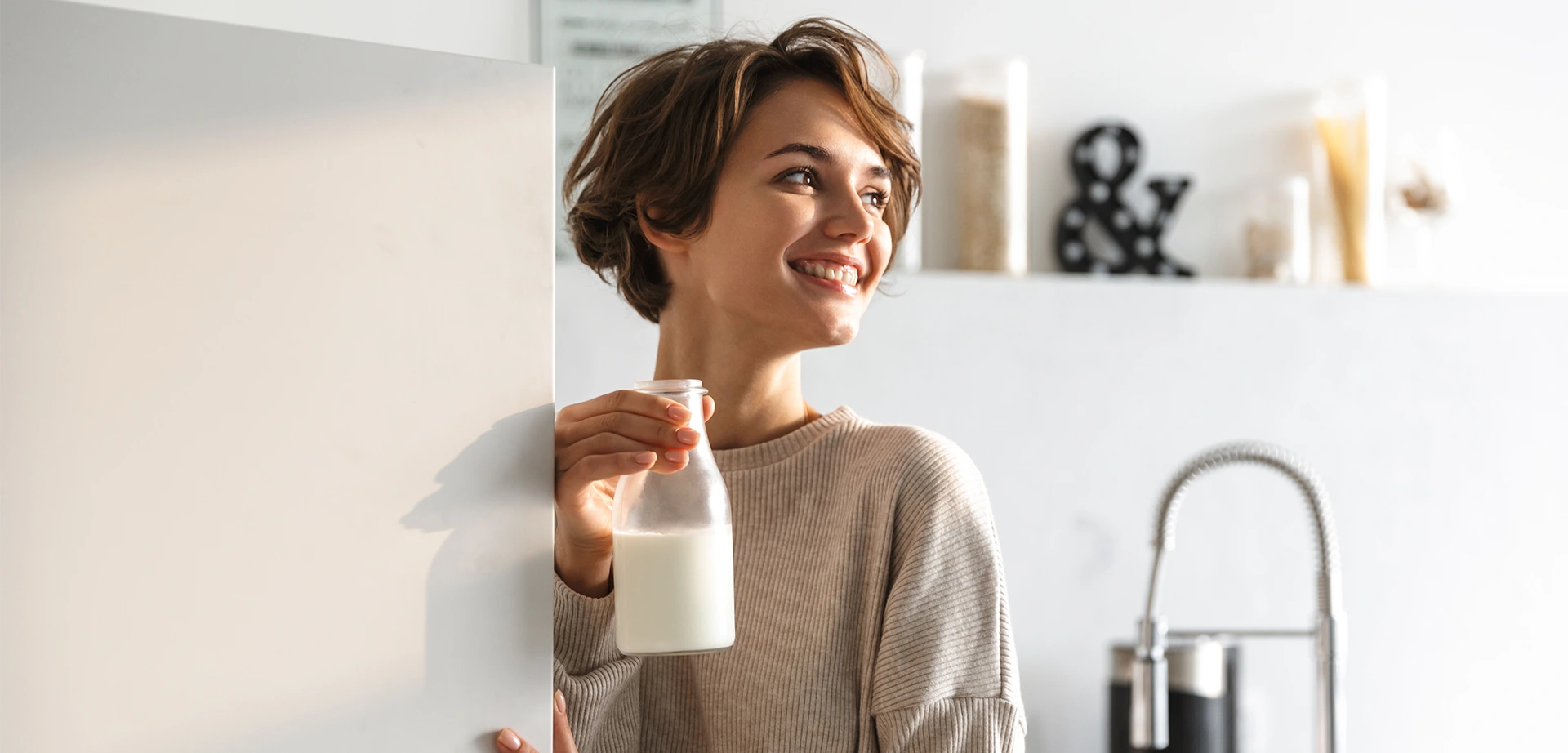 Person öffnet Kühlschranktür und hält eine Milchflasche in der anderen Hand; helle, moderne Küche im Hintergrund.