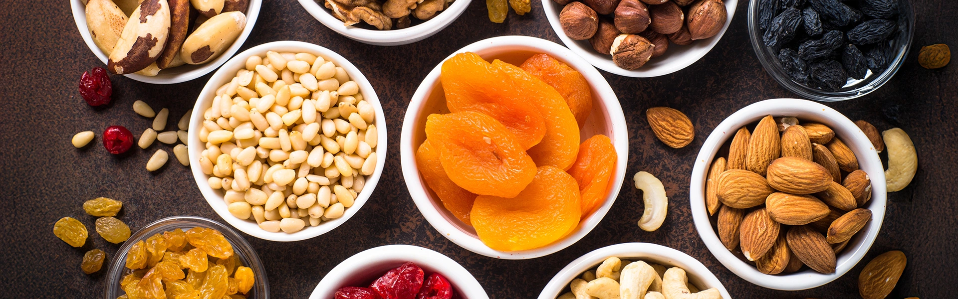 Proteinreiche Snacks Verschiedene Schälchen mit Nüssen und Trockenfrüchten auf einem Tisch, nebeneinander auf dunklem Hintergrund angeordnet.