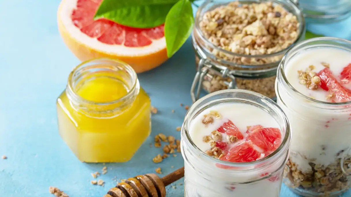 Gesunde, proteinreiche Snacks Zwei Müsligläser mit Joghurt, Granola, Nüssen und Blutorange, daneben ein Glas Honig, alles auf einem hellblauen Hintergrund.