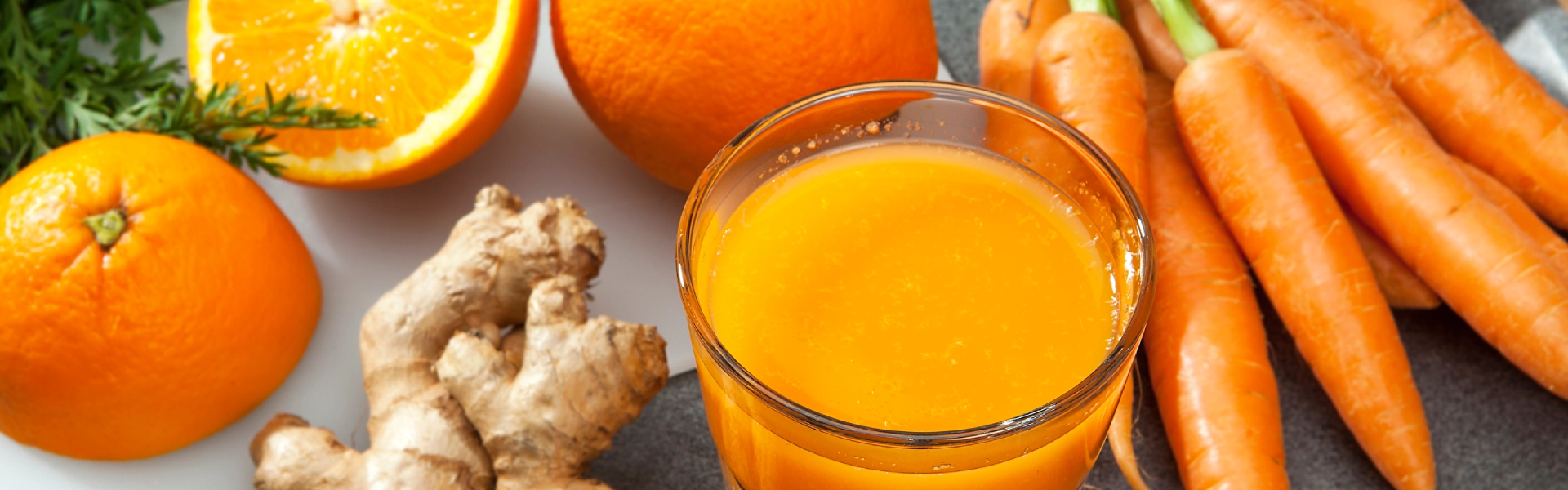 Ein Glas voll orangenem Saft steht auf einem Tisch. Daneben liegen eine große Ingwerknolle, zwei Orangen sowie Möhren.