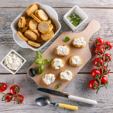 Kleine Häppchen aus Baguette und Hüttenkäse wurden auf einem Brettchen angerichtet. Daneben liegen Rispentomaten, Basilikum, weitere Baguette-Scheiben und Hüttenkäse in einer Schüssel.
