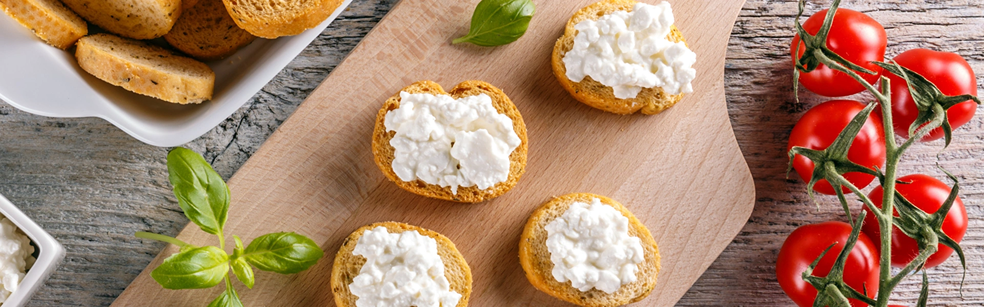 Kleine Häppchen aus Baguette und Hüttenkäse wurden auf einem Brettchen angerichtet. Daneben liegen Rispentomaten, Basilikum, weitere Baguette-Scheiben und Hüttenkäse in einer Schüssel.