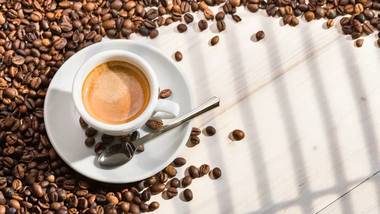 Eine Tasse Kaffee Crema steht auf einem weißen Holzuntergrund und ist umgeben von gerösteten Kaffeebohnen.