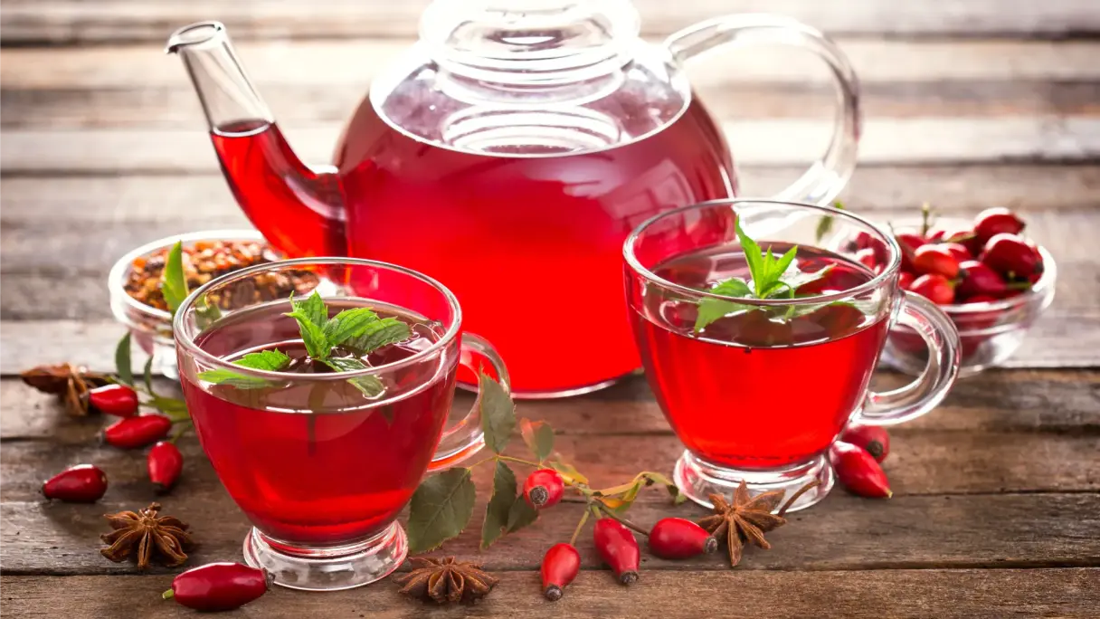 Hagebutten-Tee in gläsernen Tassen und einer Kanne. Daneben Hagebutten und Sternanis auf einem hölzernen Untergrund.