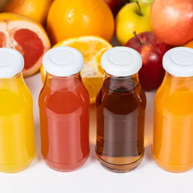 Fruchtsaft Vier Flaschen mit verschiedenfarbigen Fruchtsäften vor frischen Früchten wie Orangen, Grapefruits, Äpfeln und Kiwis – zeigt natürliche Vielfalt.