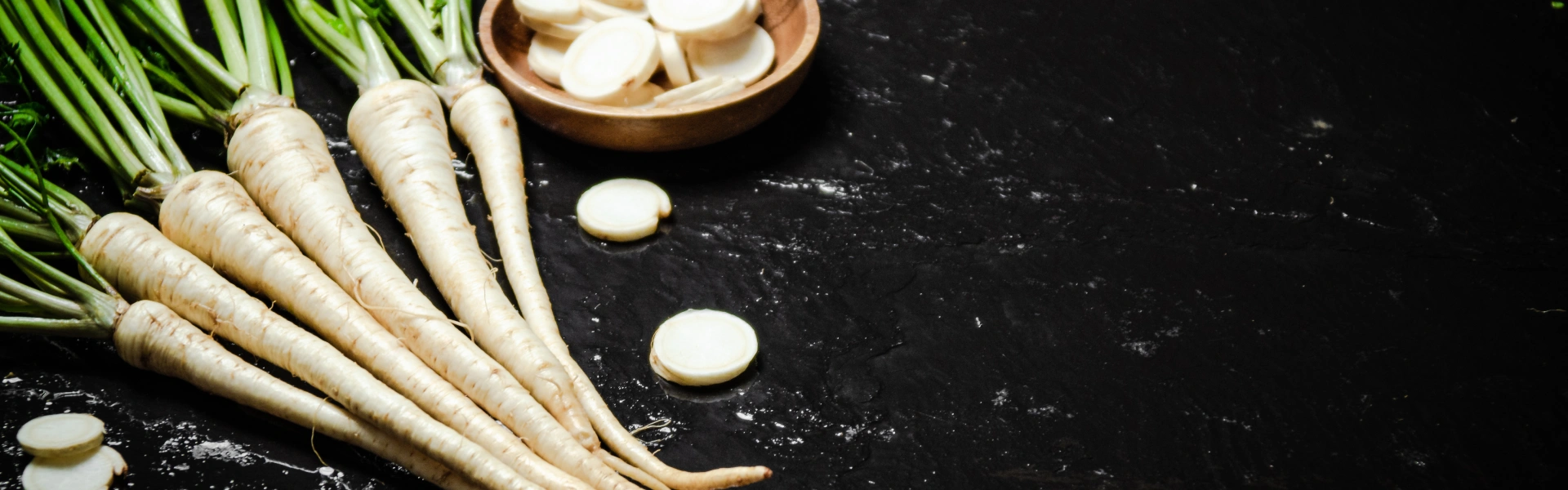 Links liegen Petersilienwurzeln mit Grün, daneben eine kleine Holzschale mit geschnittenen Scheiben, von denen einige auf der schwarzen Steinplatte verstreut sind.