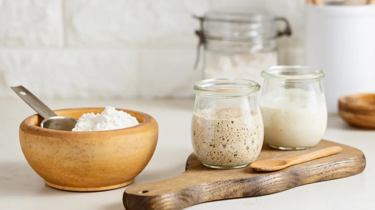 Vor weißem Hintergrund: Holzschale mit Mehl und Löffel, daneben auf Holzbrett zwei Glastöpfchen mit reifem und frischem Sauerteig.