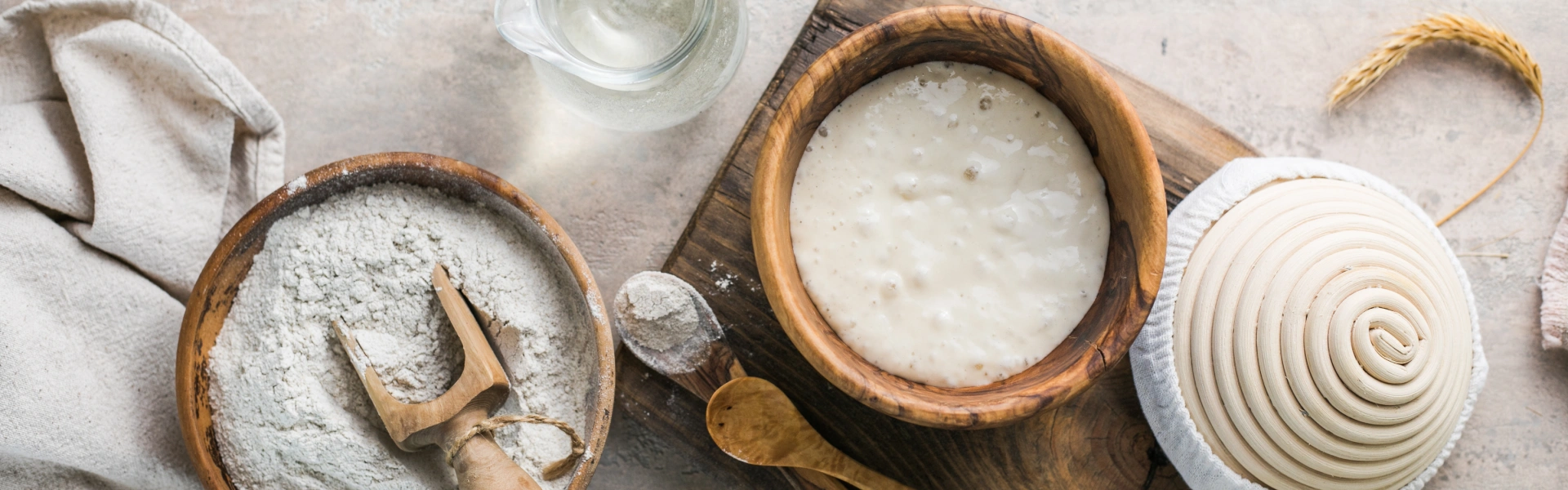 Auf hellem Untergrund: Mehlschale, daneben auf Holzbrett Schüssel mit Sauerteig, Gärkörbchen, zwei Kochlöffel und oben eine Karaffe.