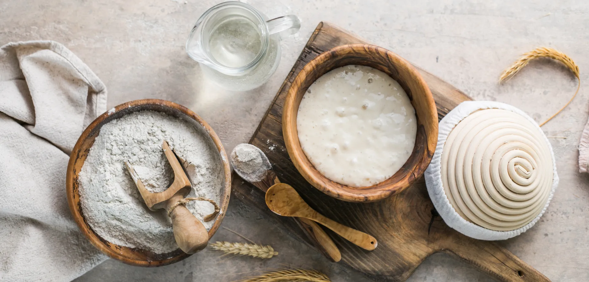 Auf hellem Untergrund: Mehlschale, daneben auf Holzbrett Schüssel mit Sauerteig, Gärkörbchen, zwei Kochlöffel und oben eine Karaffe.