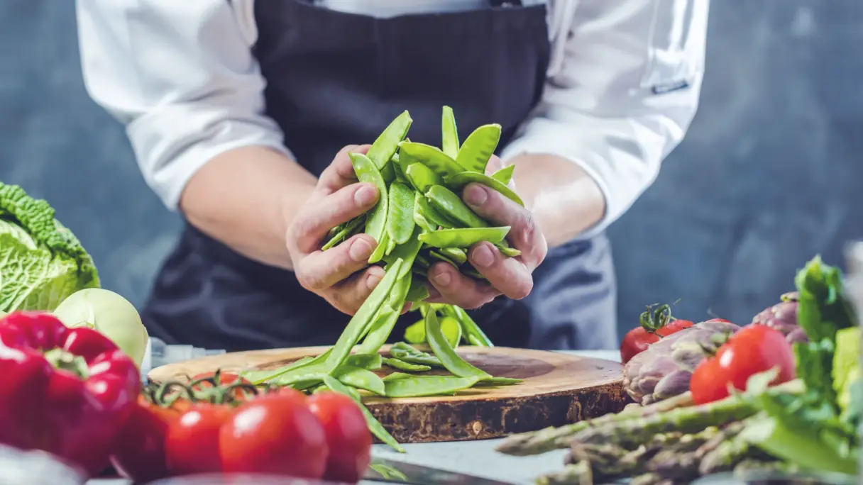 Sugar snap peas held in hands with assorted vegetables in the foreground.
