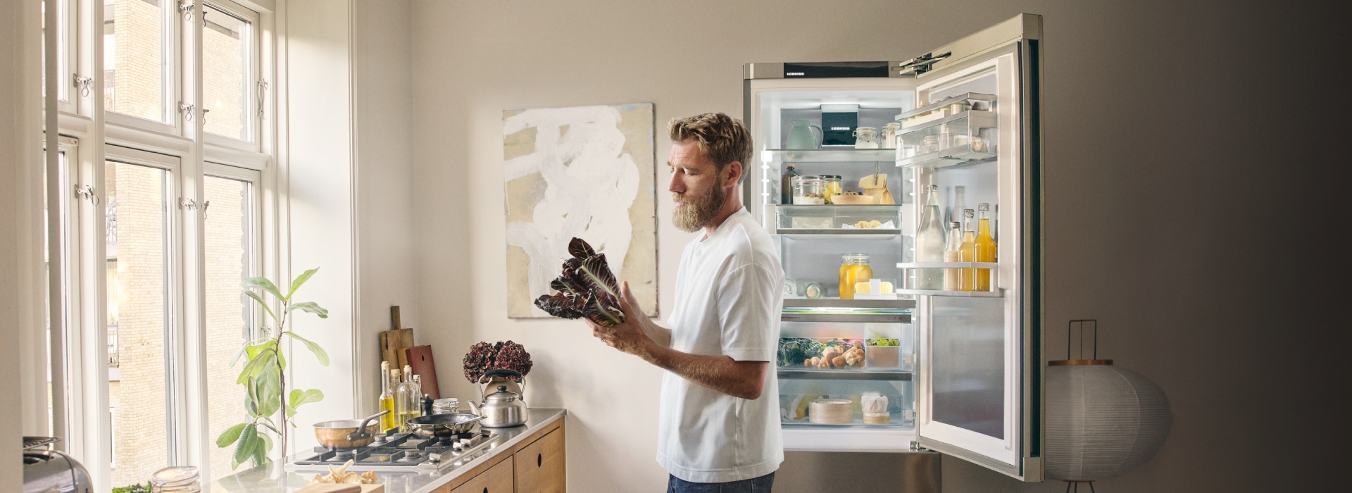 Mikkel Karstad standing in a modern kitchen holding fresh salad in front of an open refrigerator filled with food and drinks.