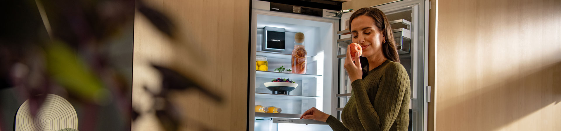 Woman standing in front of open fridge, sniffing a peach