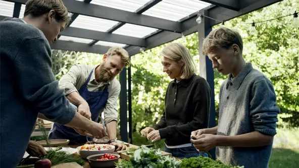 People preparing fresh ingredients together on a table under a covered terrace surrounded by greenery.
