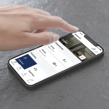 Hand using a smartphone with the SmartDevice app; the display shows temperature zones and functions of a combined fridge‑freezer.