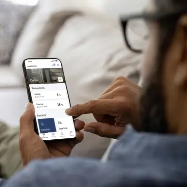 Person using the SmartDevice app on a smartphone; the screen shows settings of a fridge‑freezer connected via the SmartDeviceBox.