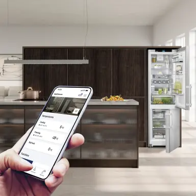 Modern kitchen with an open fridge‑freezer; a hand holds a smartphone with the SmartDevice app for temperature control.