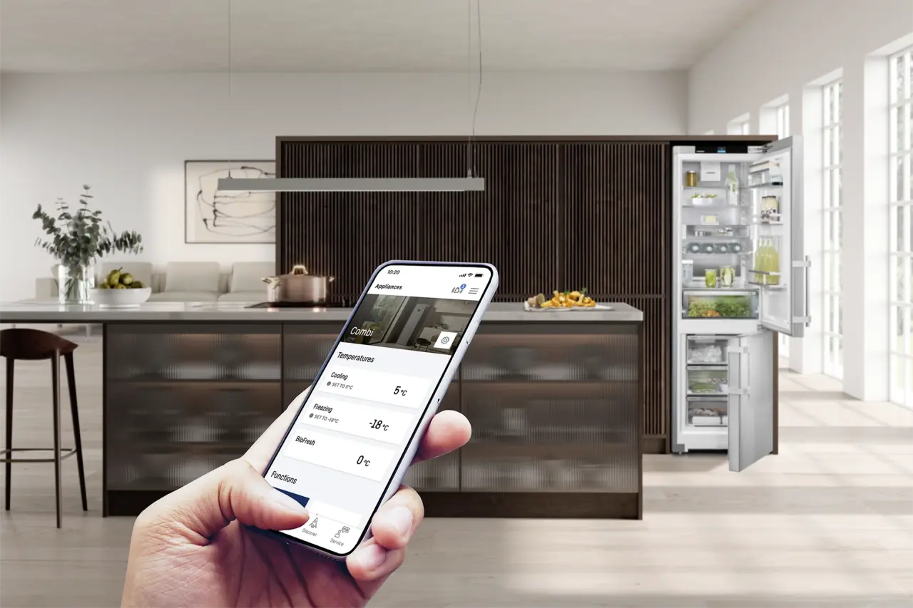 Modern kitchen with an open fridge‑freezer; a hand holds a smartphone with the SmartDevice app for temperature control.