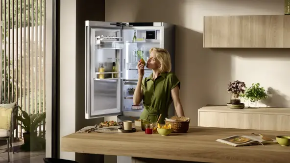 Woman smelling a pear in a light, wooden kitchen and a freestanding open fridge in background