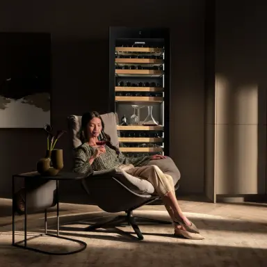 Woman sitting in in lounge chair and smelling her wine glass with wine fridge in the background