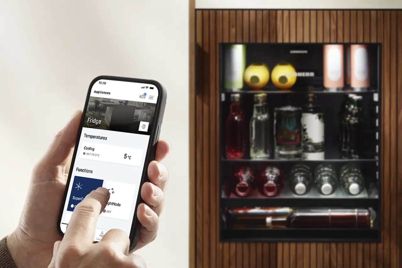 Person hält Smartphone, tippt auf Display, unscharf im Hintergrund Unterbau-Getränkekühlschrank mit Glastür