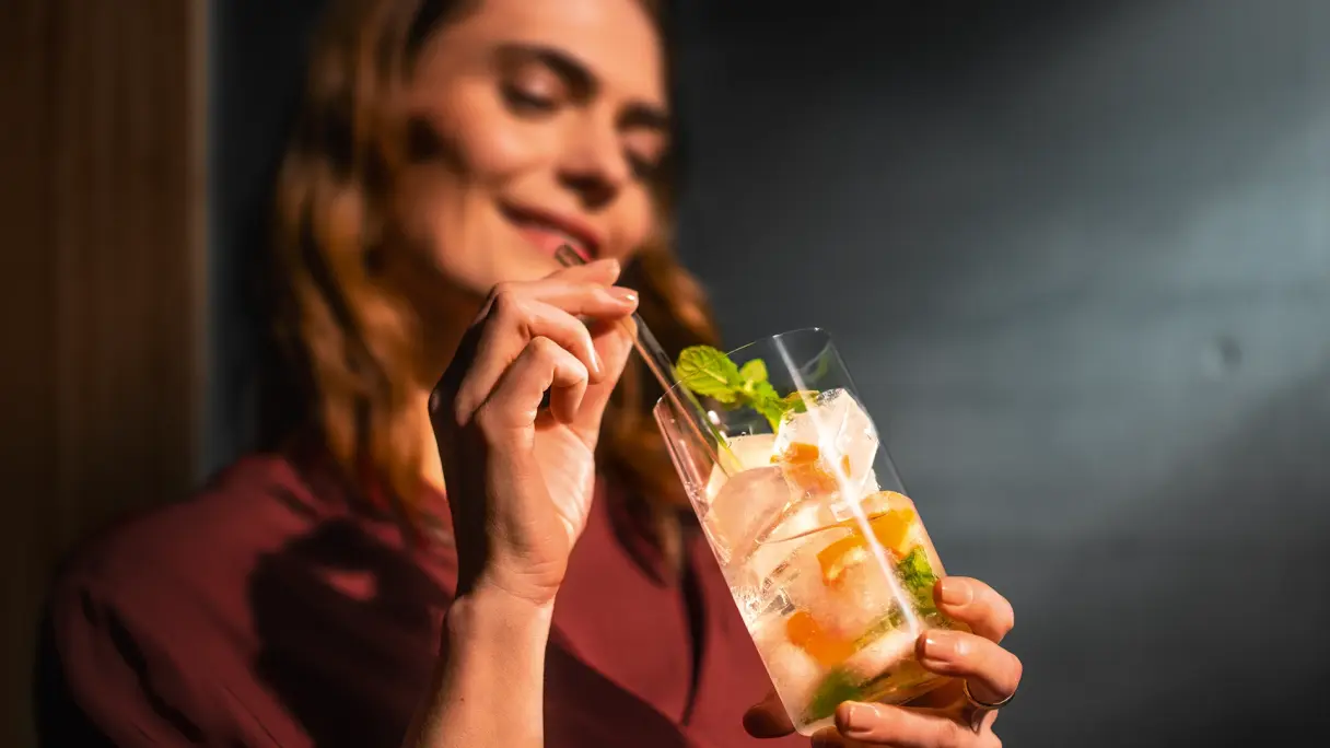 Person hält Drink mit Zitrus und Minze in der Hand, unscharfer Hintergrund im Innenraum.