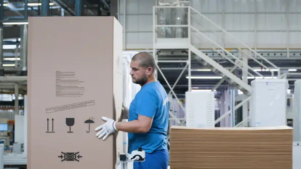 In a factory hall, a man is packaging a fridge using cardboard and ensuring the fridge is securely wrapped for protection
