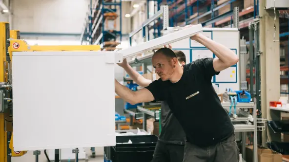 Two men are working together to assemble the door of a freezer in a spacious factory hall. They are focused on their task to ensure a precise fit.