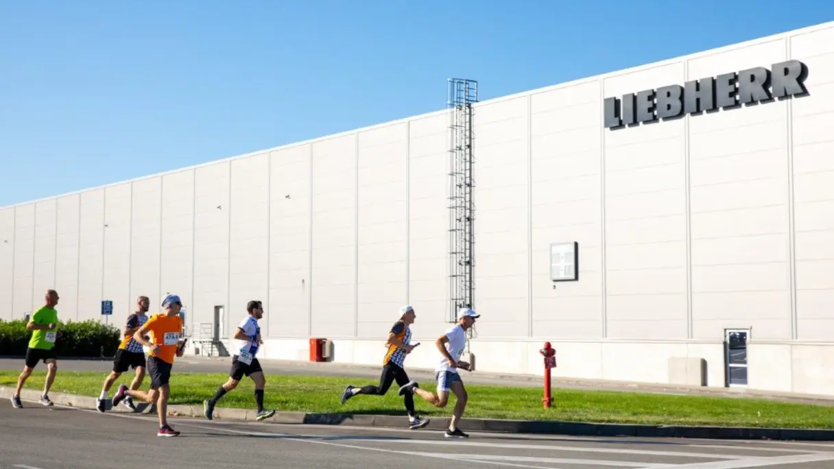 A group of runners is seen jogging near a Liebherr building. The vibrant scene showcases individuals of various backgrounds enjoying the outdoors.
