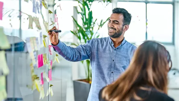  A cheerful man stands in an office environment, actively writing on a whiteboard filled with colorful post-its. His positive demeanor reflects enthusiasm and creativity as he shares ideas with colleagues.