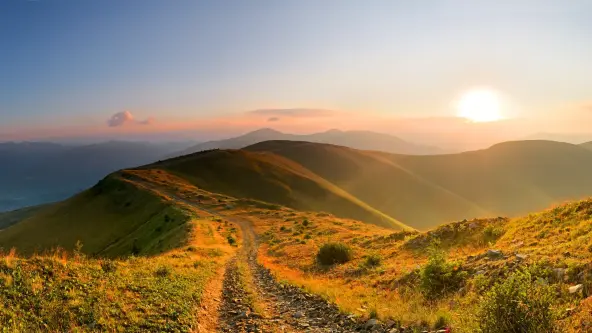 A serene view of a soft mountain range at sundown, with a winding path along the ridge. The sky is painted in warm hues of orange and blue, casting a gentle glow over the landscape.
