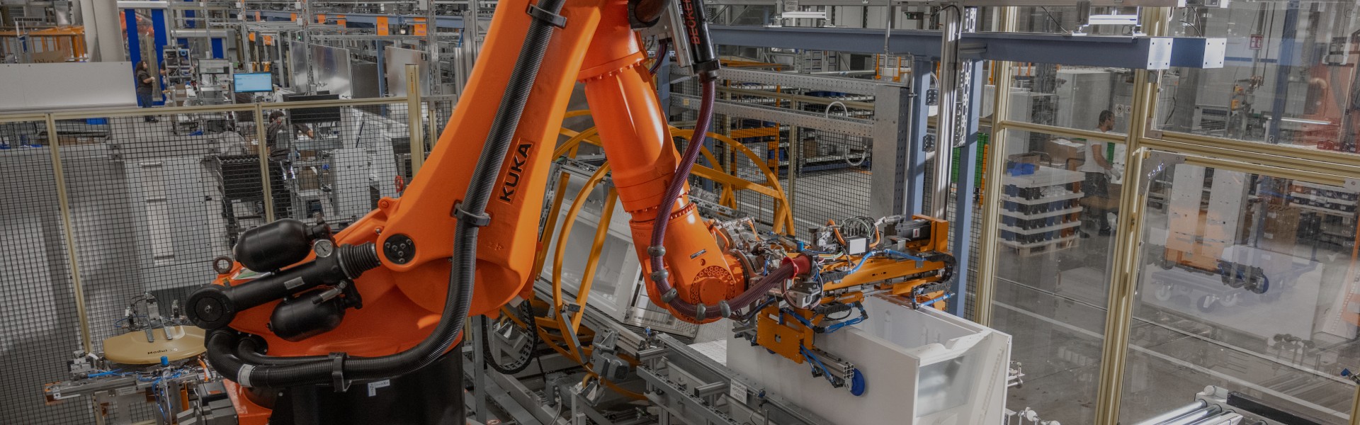 In a spacious factory hall, an orange robotic arm is grabbing a fridge from the conveyor belt. 