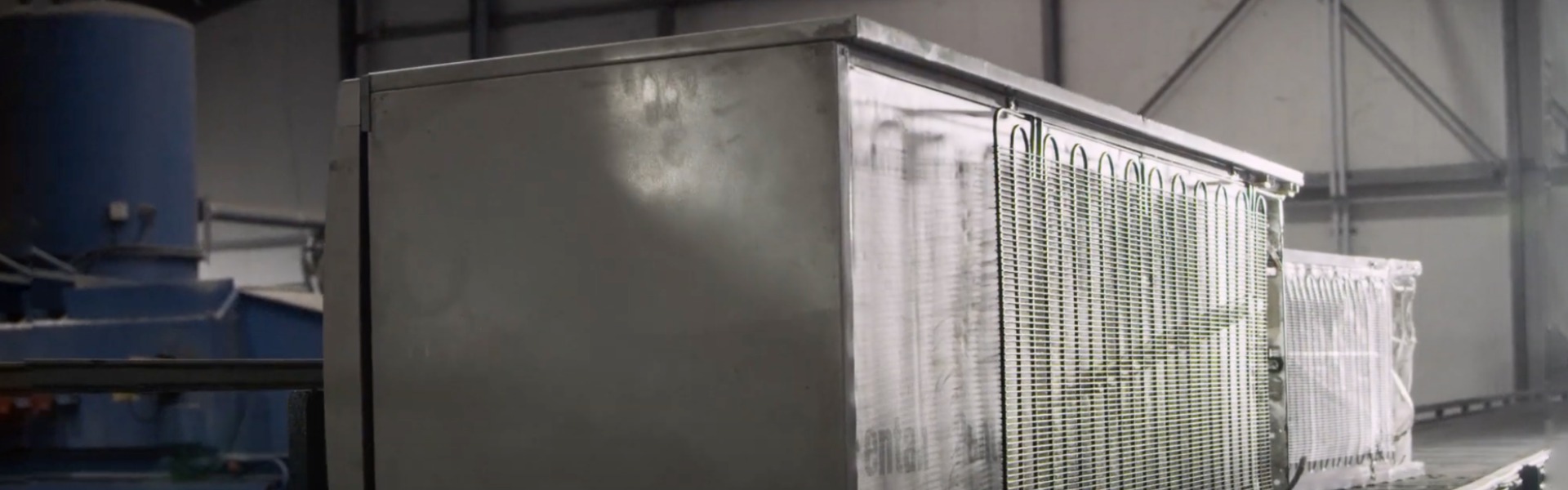 An old and stained fridge lies with the back visibile on a conveyor belt within an industrial hall. Behind you can see another fridge on the line.