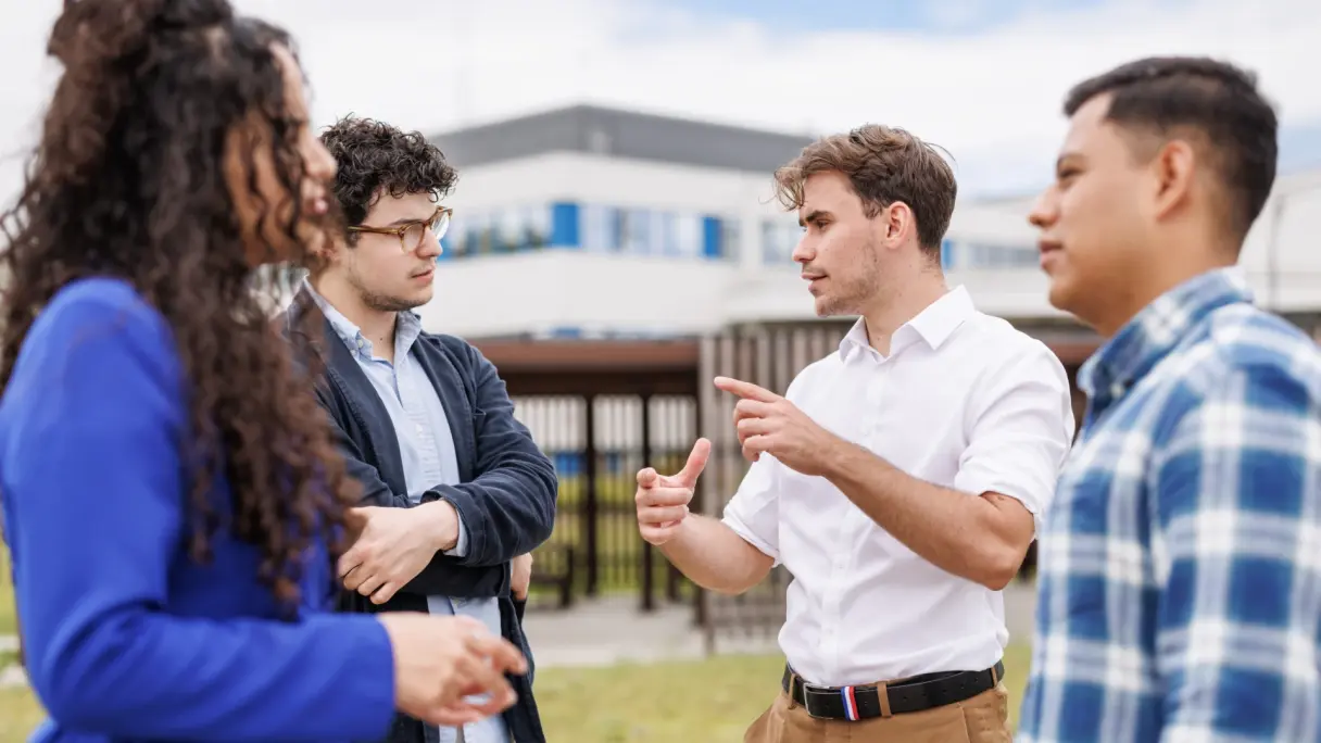 A diverse group of students is gathered outside, animatedly talking and using gestures to emphasize their points. 