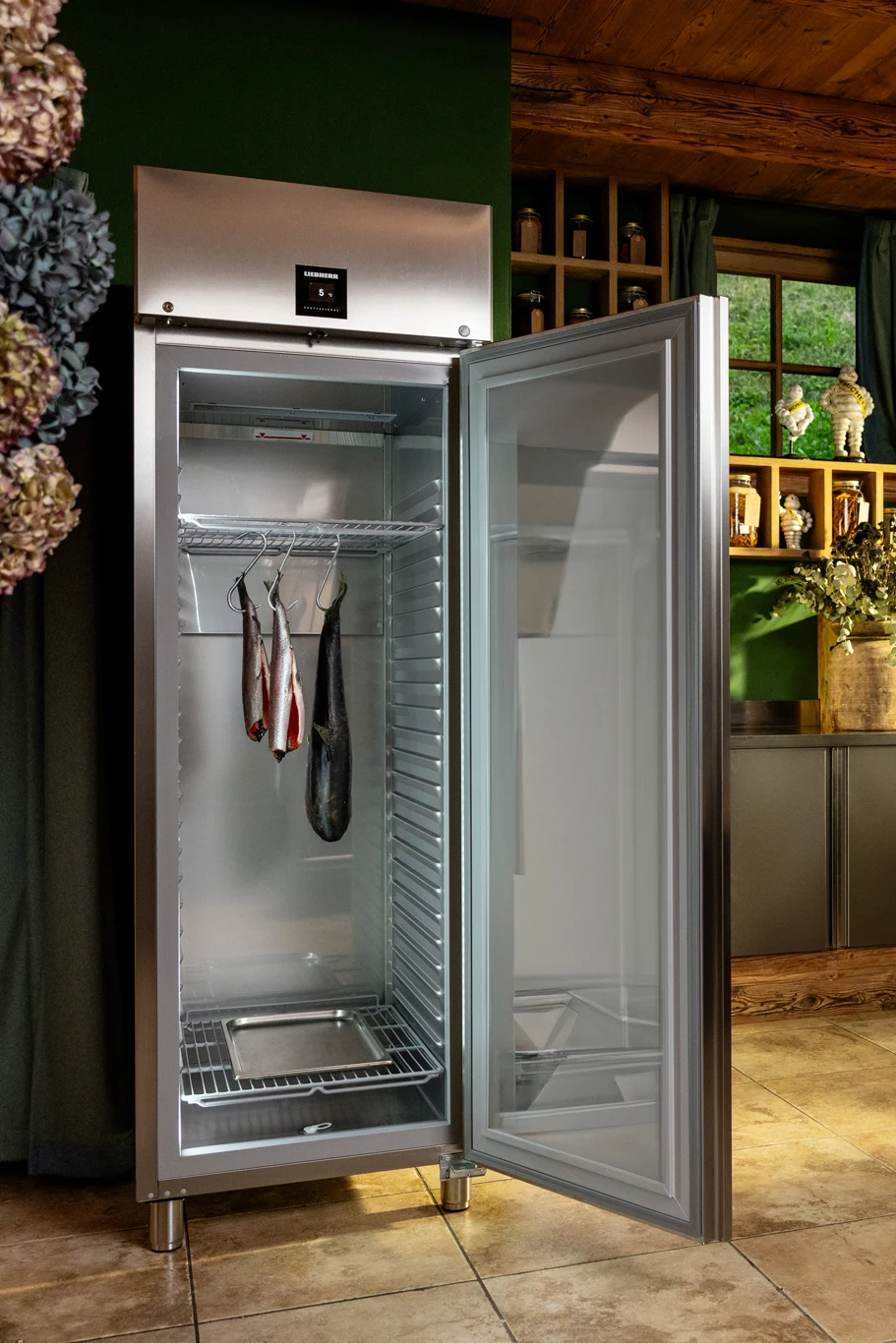 Food Service fridge with open door in a rustic professional kitchen The Food Service fridge stands with its doors open, showcasing fresh fish hanging inside in an appealing display. The rustic professional kitchen features open shelves filled with various ingredients, while a wooden table and chairs create an inviting atmosphere.