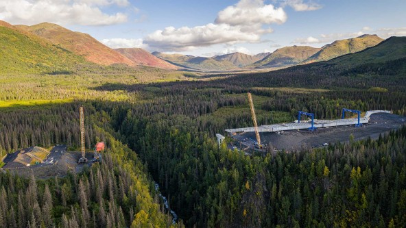 Juneau Creek Bridge construction near Cooper Landing, Alaska, captured from above with crawler crane activity and staged bridge framework in a remote mountain landscape.