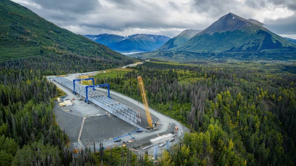 Drone view of construction for Alaska’s 951‑foot Juneau Creek Bridge near Cooper Landing, with a Liebherr LR 1300.1 SX crawler crane working beside staged steel bridge sections in a remote valley.