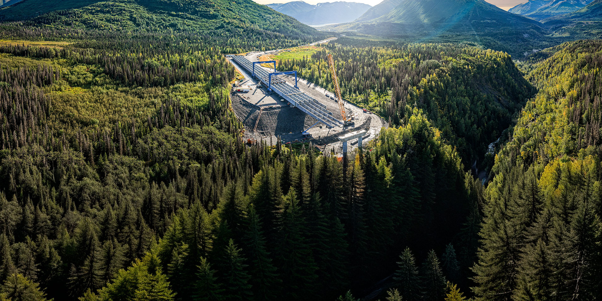 Wide drone panorama of a bridge construction site along a winding road through dense forest, framed by mountains and glowing evening light.
