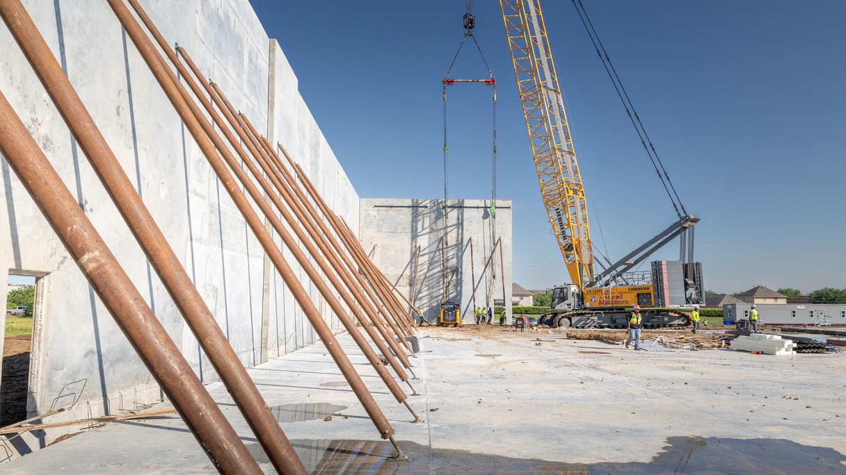 Ground-level view of a construction site where a Liebherr crawler crane positions a precast concrete wall panel